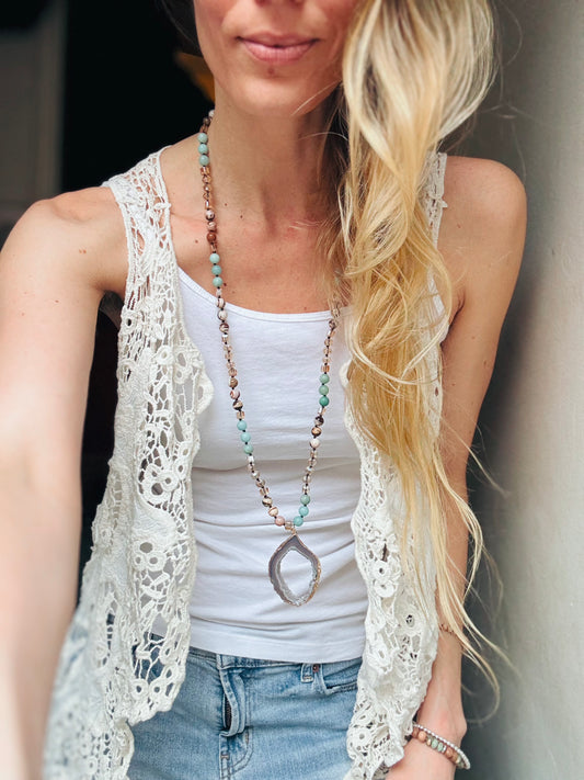 A boho-style necklace named The Odessa Necklace, inspired by the ocean’s rhythmic waves and serenity. It features soothing Amazonite beads, earthy Sandstone, and radiant gold glass beads, creating a harmonious blend of land and sea. At its center, a shimmering Druzy Agate pendant reflects the beauty of sunlit tide pools and hidden ocean treasures. A symbol of tranquility and strength, this handcrafted piece embodies the ebb and flow of nature.