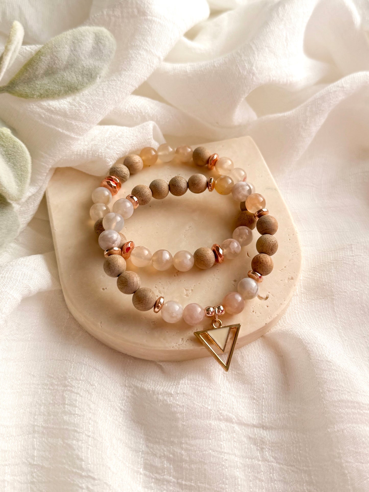 A delicate gemstone bracelet featuring soft pink and white Flower Agate beads, warm Matte Beechwood accents, and a triangle charm. The design is inspired by cherry blossoms, symbolizing renewal and fleeting beauty. The bracelet embodies growth, transformation, and harmony with nature.