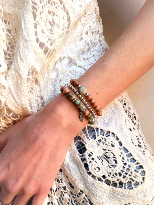 A handcrafted bracelet set featuring Serpentine Jasper beads in earthy green tones, symbolizing transformation and inner peace. Warm, natural Rosewood beads provide grounding energy, while a delicate feather charm dangles, representing freedom and intuition. Designed for free spirits and wanderers, this set embodies balance, resilience, and adventure.