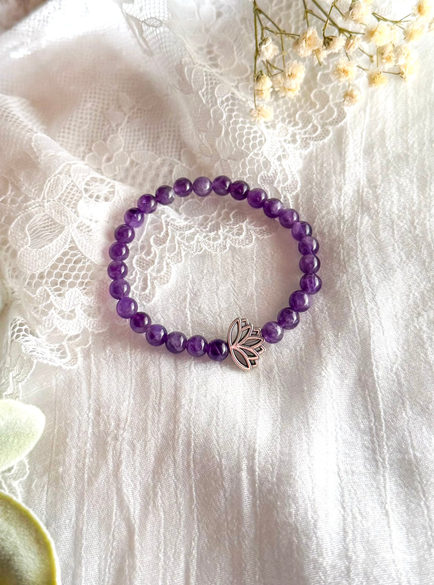 A gemstone healing bracelet made with smooth 6mm purple Amethyst beads and a single silver-toned Lotus charm, symbolizing spiritual growth and inner peace. The bracelet is arranged in a circle on a soft, neutral background.