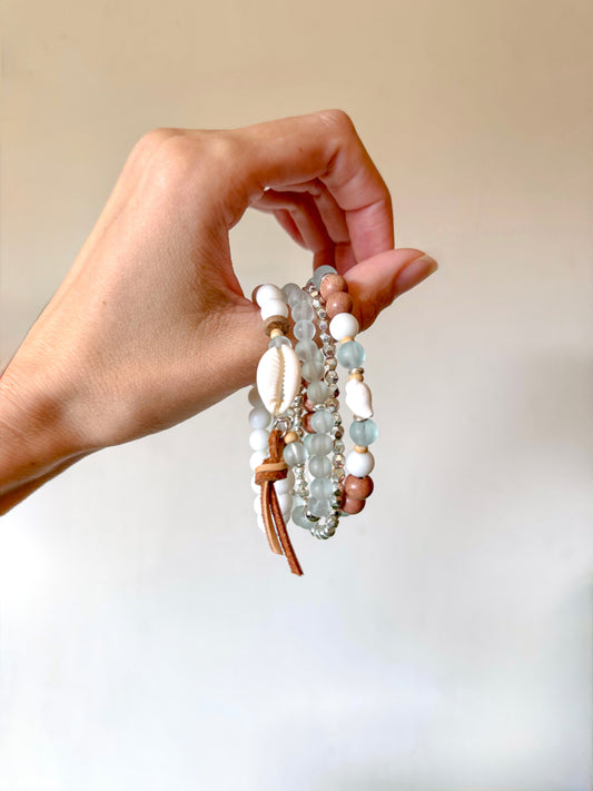 A coastal-inspired bracelet set featuring smooth, frosted sea glass beads in soft ocean hues and a delicate conch shell charm. The bracelets are styled on a sandy, beach-themed background with seashells and sunlight, evoking healing, intuition, and laid-back summer energy.