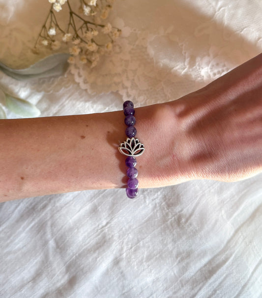 A gemstone healing bracelet made with smooth 6mm purple Amethyst beads and a single silver-toned Lotus charm, symbolizing spiritual growth and inner peace. The bracelet is arranged in a circle on a soft, neutral background.