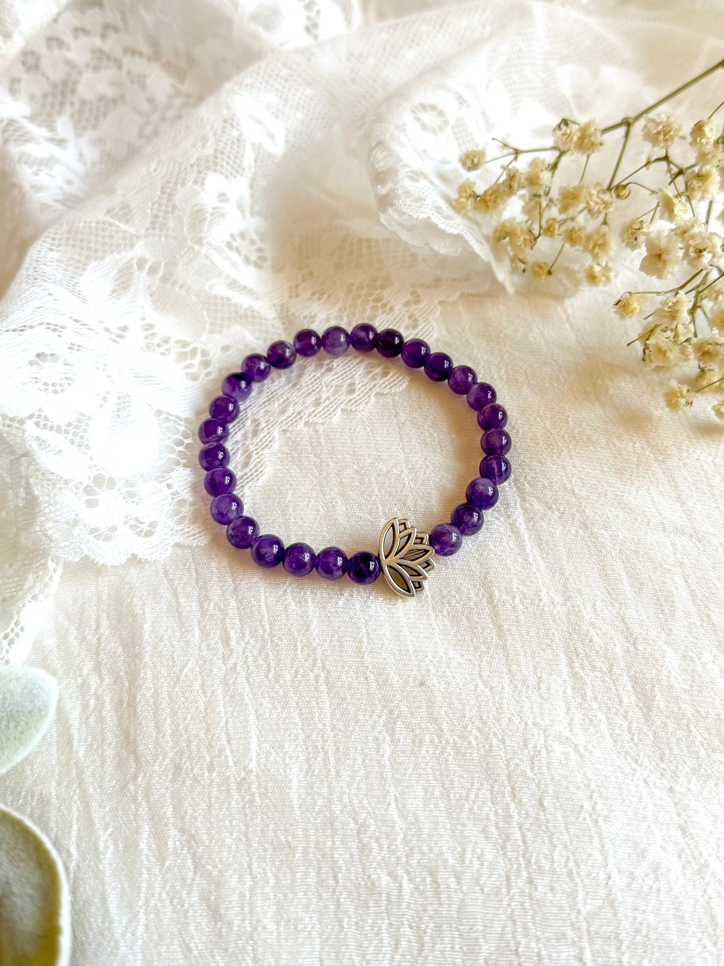 A gemstone healing bracelet made with smooth 6mm purple Amethyst beads and a single silver-toned Lotus charm, symbolizing spiritual growth and inner peace. The bracelet is arranged in a circle on a soft, neutral background.