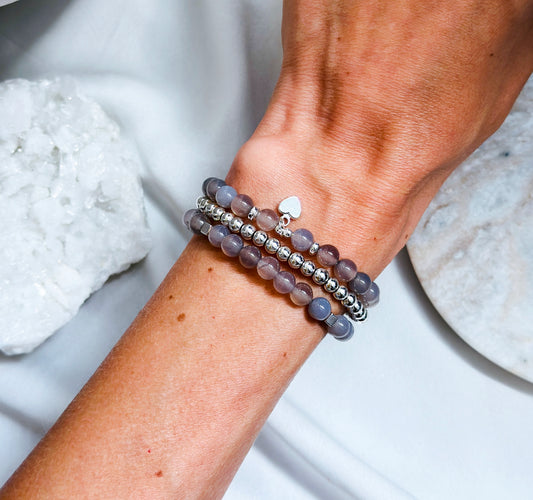 A set of Grey Agate gemstone bracelets with silver heart charms, elegantly designed to promote tranquility and self-love. The bracelets feature smooth, polished Grey Agate beads with soft, calming hues and natural patterns. Each bracelet is accented with a delicate silver heart charm, symbolizing love, self-acceptance, and emotional harmony. The set is arranged beautifully, highlighting its soothing and elegant aesthetic.