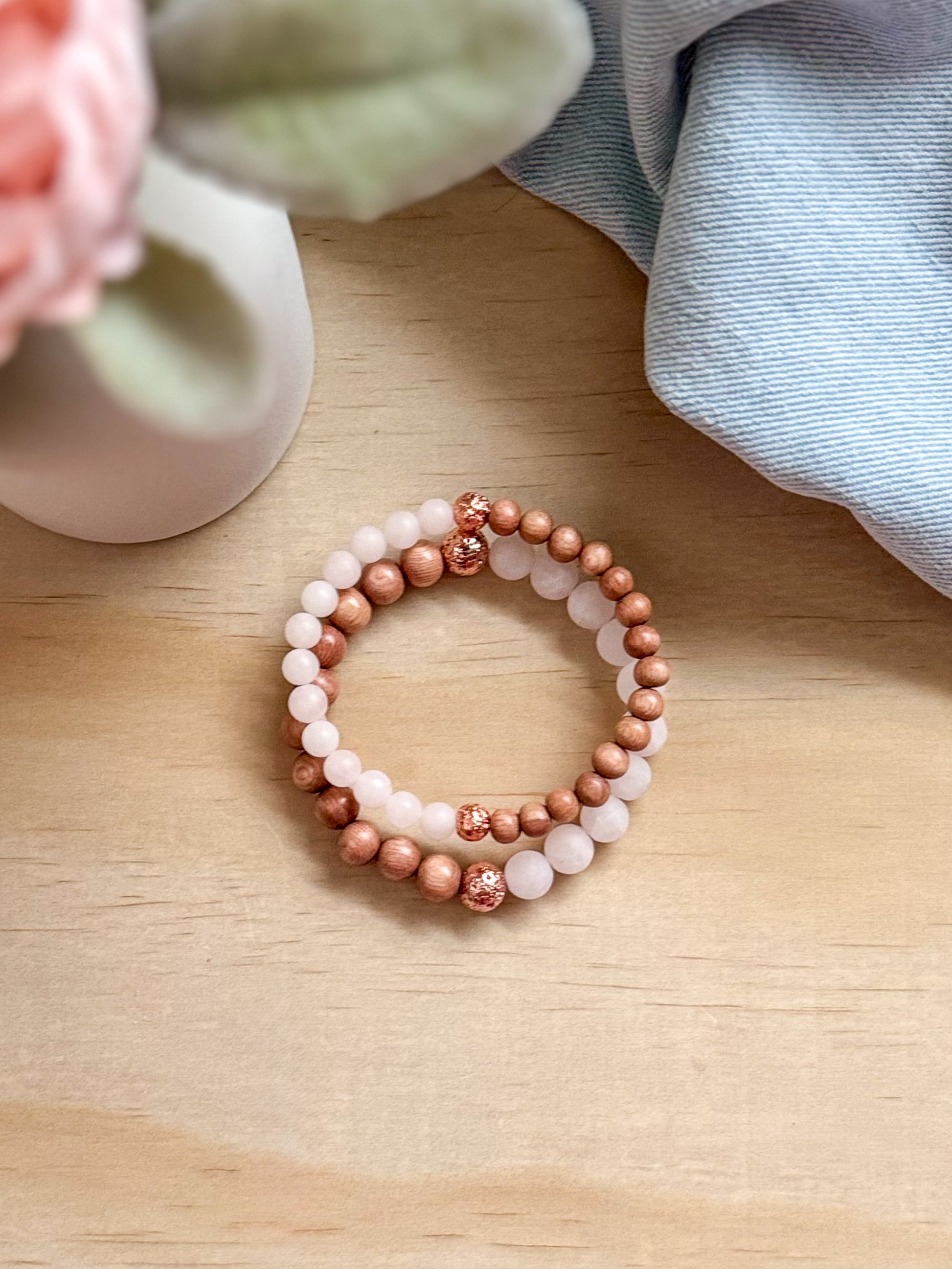 A handcrafted bracelet featuring matte, earthy brown Rosewood beads paired with soft, translucent pink Rose Quartz stones. The bracelet is displayed against a natural, minimalist background with a small green sprig, highlighting the contrast between the wood and the crystals.