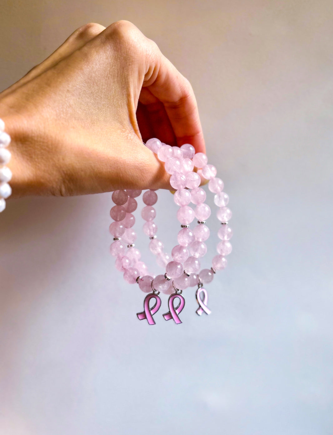 Hand holding pink beaded bracelets with breast cancer awareness charms against a light background