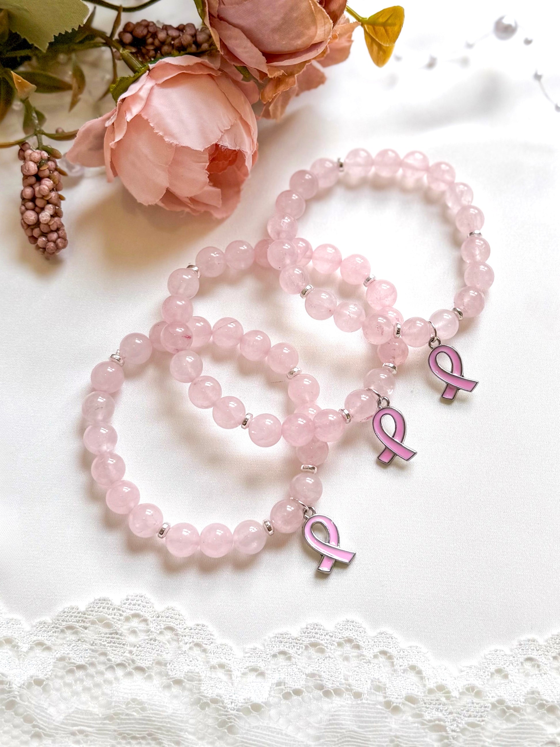 Pink beaded bracelets with breast cancer awareness ribbons on a white surface with flowers.