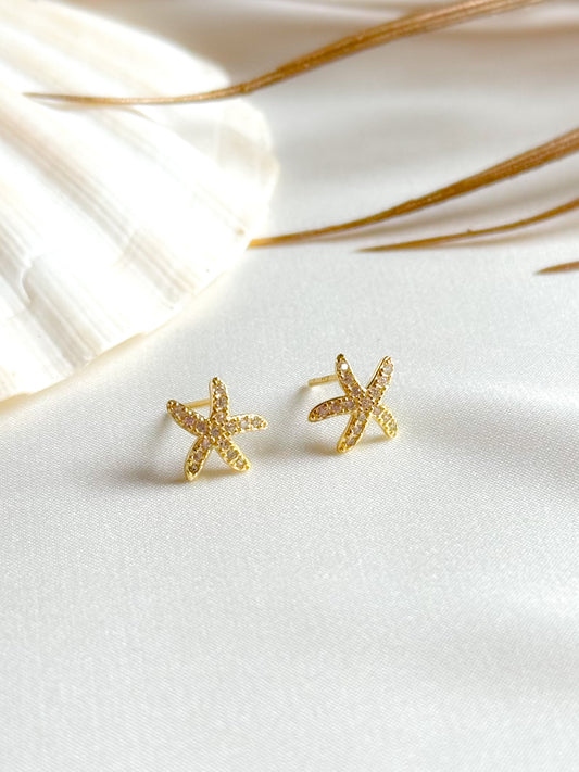 A pair of delicate stud earrings inspired by the ocean, featuring 14k gold and sparkling cubic zirconia. Styled against a light, summery backdrop, the earrings are minimal, elegant, and perfect for beach days or everyday wear. Text overlay highlights their ocean-inspired design and summer-ready sparkle.