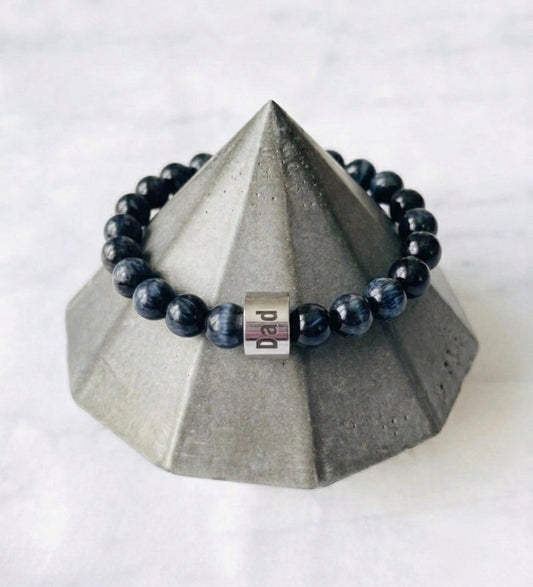 Black beaded bracelet with a silver dad bead for Father’s Day  on a geometric gray stand against a white background
