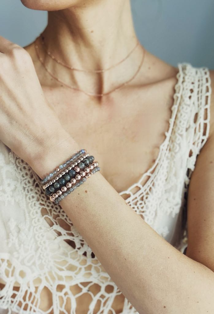 Lifestyle shot of a person wearing a stack of five delicate Little Lotus gemstone healing bracelets in shades of grey, blue, and rose gold, paired with a layered gold tarnish proof necklace and a white crochet lace top. Made in toronto ontario Canada